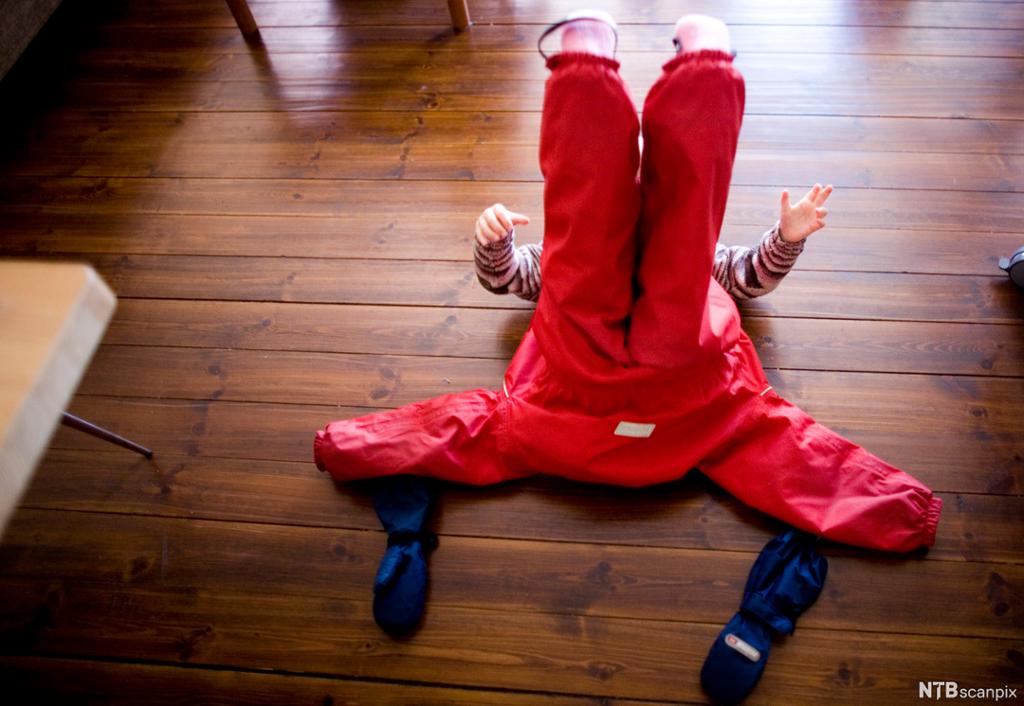Et barn ligger på gulvet og strekker beina i taket. Har utedress på beina. Votter ligger ved siden. Foto.