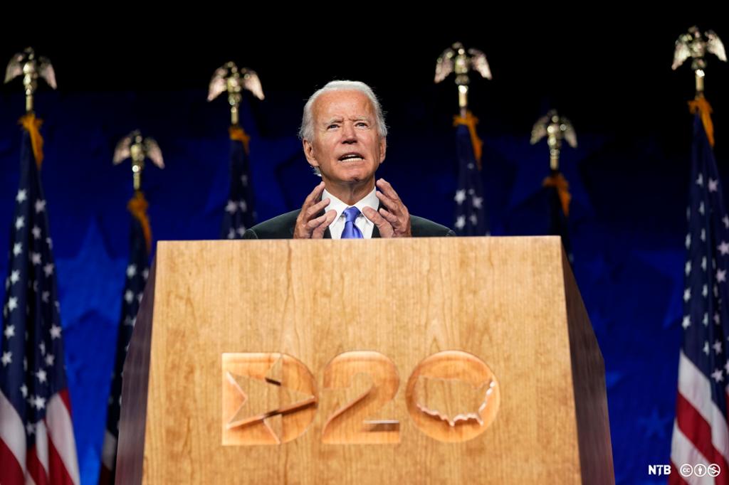 Photo: We see Joe Biden standing behind a wood pulpit with 'D20' engraved on it. Behind Biden, there are six American flags, each with a metal eagle on top. Joe Biden is an elderly, white-haired man, wearing a suit and a blue tie. 