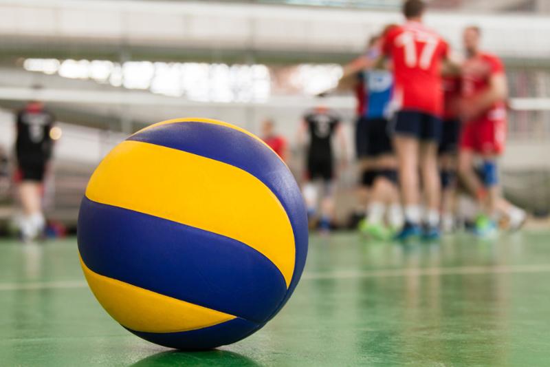 Gul og blå volleyball som ligger i ro på grønt gulv. Volleyballspillere og volleyballnett vises diffust i bakgrunnen. Bilde.