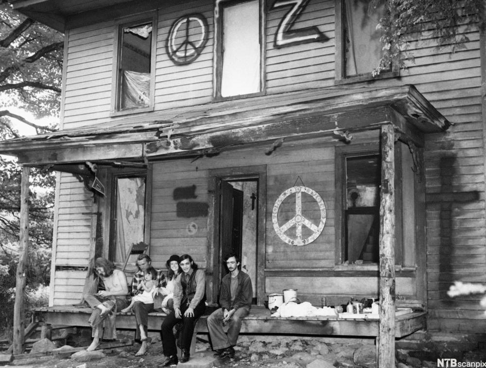 Fem voksne hippier sitter på en terrasse utenfor et hus som er dekorert med freds-symboler. To av dem har barn på fanget. På terrassen står det malingsspann og annet utstyr. Svart-hvitt-foto. 