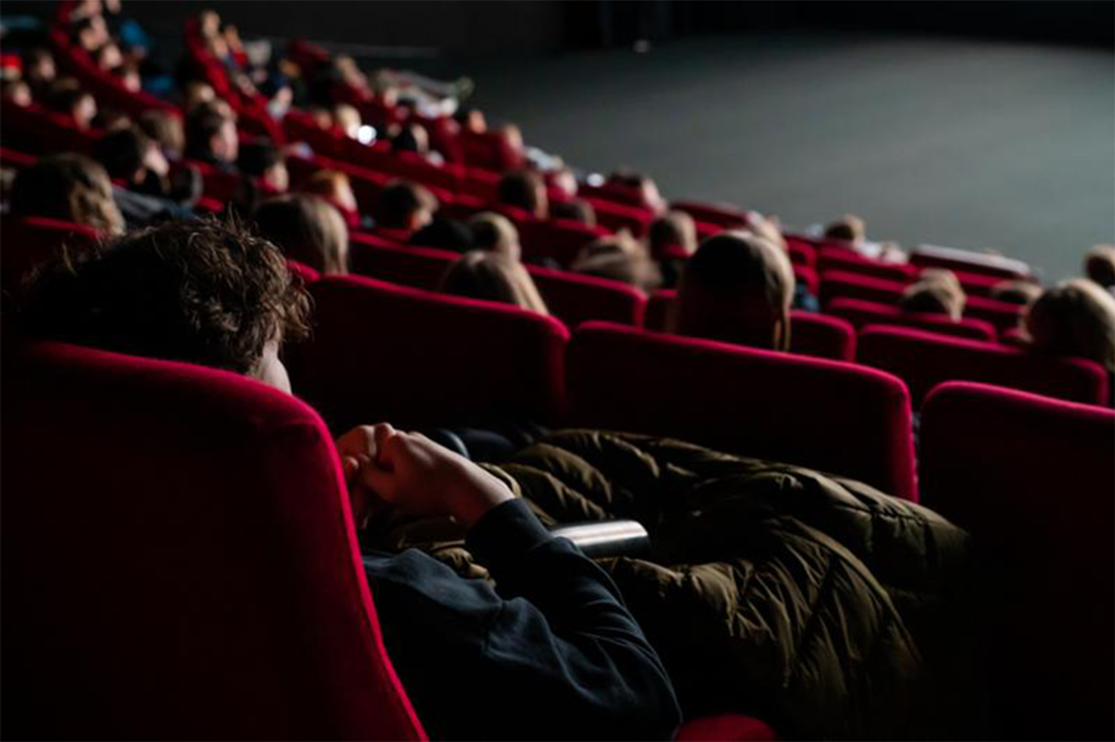 Røde stolrader i en kinosal sett bakfra. Hodene til de som sitter i stolene, lyses opp av lyset fra kinolerretet. Foto. 