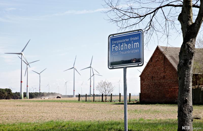Et landlig område. Til høyre en rødmalt låve, i bakgrunnen en rekke mindre vindmøller. I forgrunnen et blått stedsskilt med stedsnavnet "Feldheim". Foto.
