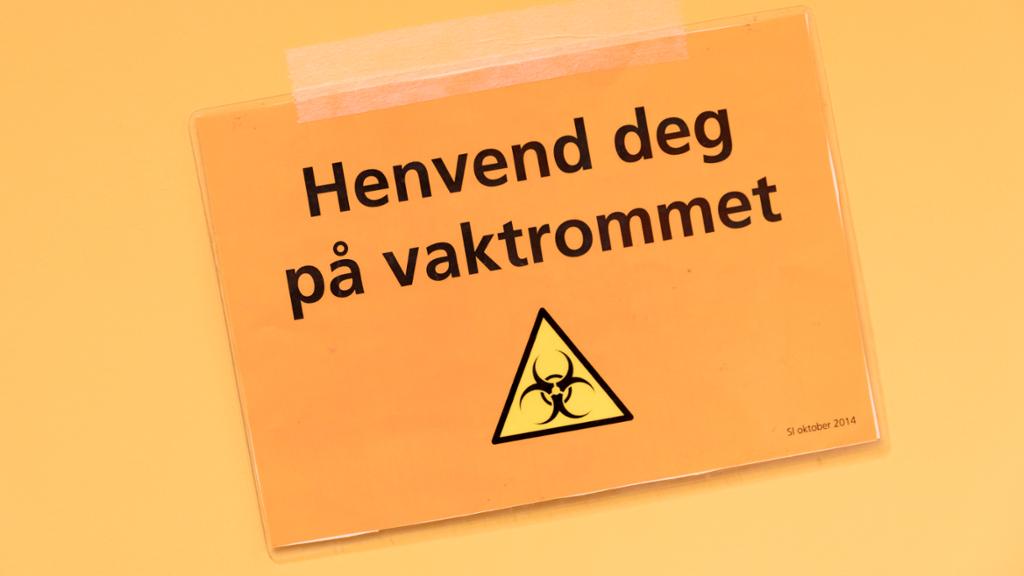 Skilt med teksten "Henvend deg på vaktrommet" og eit gult og svart fareskilt med symbol for biologisk fare. Foto.