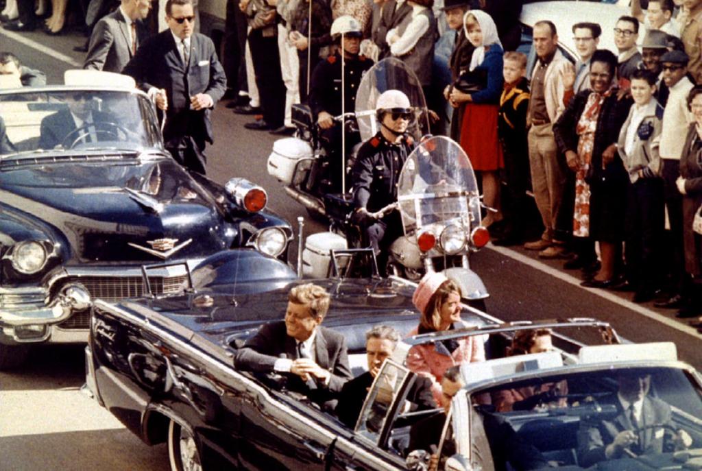 President John F. Kennedy blei drepen av ein snikskyttar under eit besøk i Dallas, Texas, 22. november 1963 . Seks vaksne personar sit i ein open bil. Dei køyrer i ein kortesje. To sikkerheitsvakter på motorsykkel køyrer òg i kortesjen. Det står folk langs heile gata. Foto.