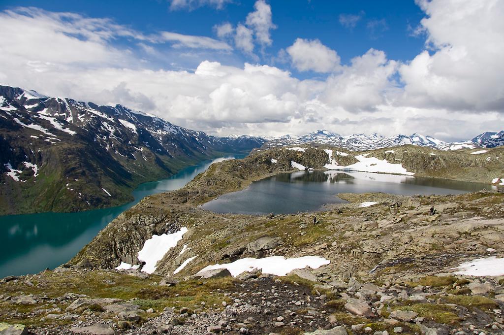 Rundt en innsjø oppe på fjellet ligger det flekker med snø. Bak ser vi enda en innsjø og snødekkede fjelltopper under en delvis skyet himmel. Foto