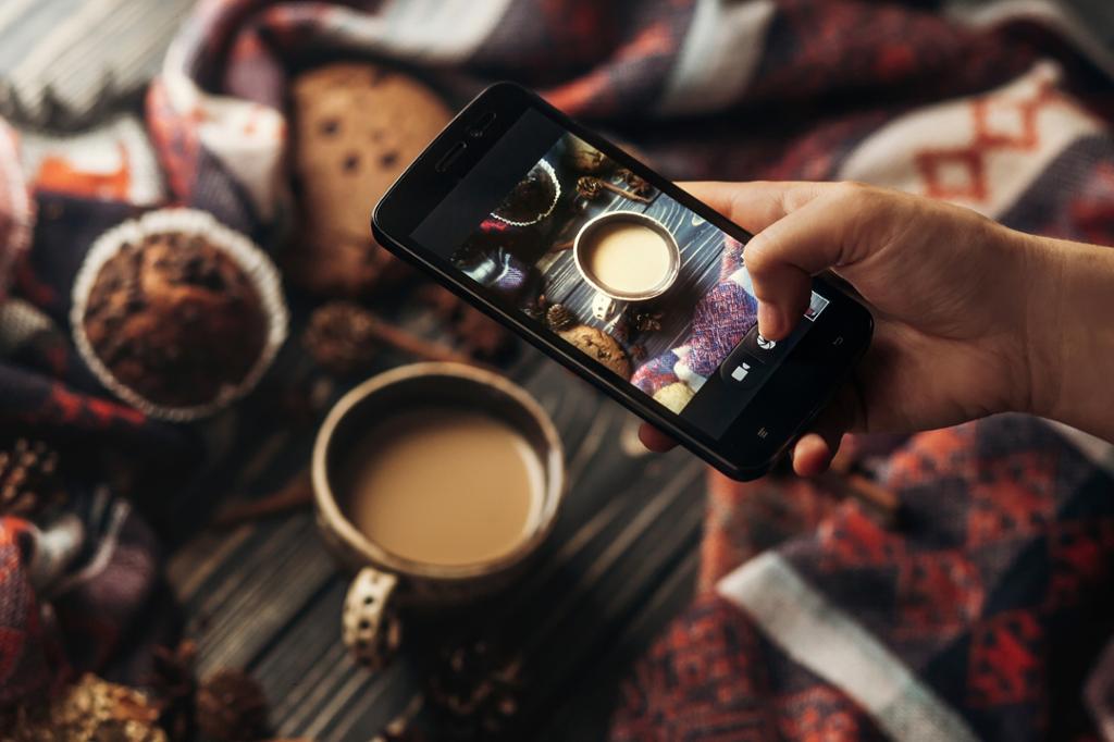 En hånd holder en telefon over et stylet oppsett med kaffe og muffins og tar et bilde. Foto.
