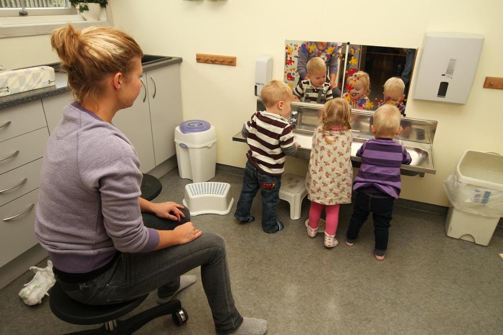 En voksen sitter på stol og ser på tre småbarn som vasker hendene. Foto.