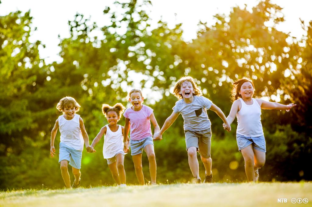 Fem barn på rekke. De holder hverandre i hendene mens de smiler og løper i en park. Foto.