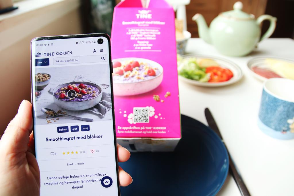 En mobilskjerm viser nettsiden til Tine Kjøkken og oppskriften på smoothiegrøt med blåbær. I bakgrunnen står en melkekartong med QR-kode på et frokostbord. Foto.