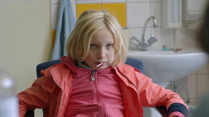 Ei niårig jente i rosa antrekk og med kjærlighet på pinne i munnen sitter på en blå stol. Hun ser skeptisk på personen overfor henne. I bakgrunnen skimtes en vask. Stillbilde fra filmen "Systemsprengeren".