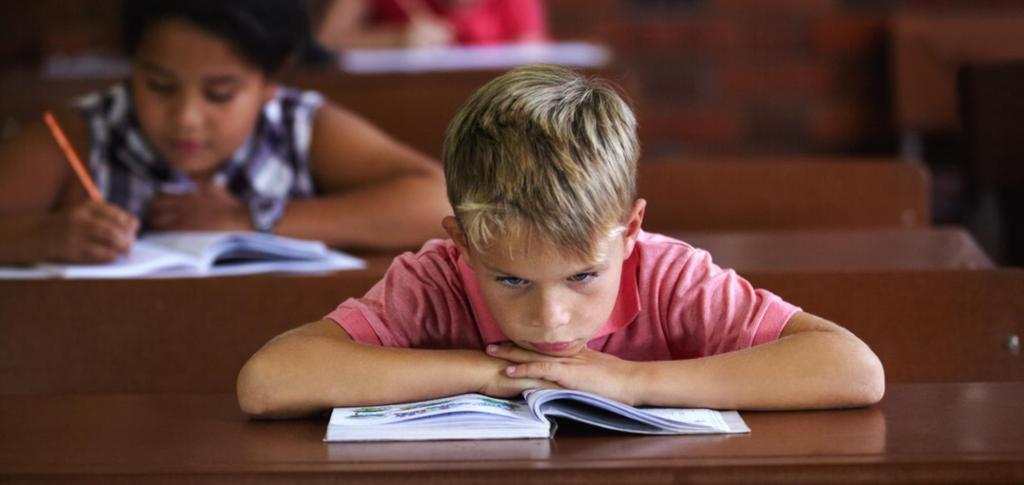 A young boy is sitting by a school desk, looking frustrated and bored. There are two other students in the background, working. Photo.