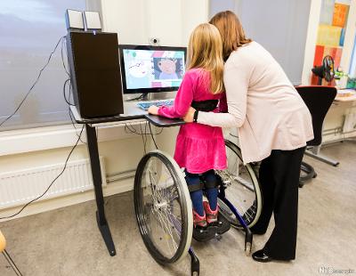 Voksen og barn med stående rullestol ved datamaskin. Foto.