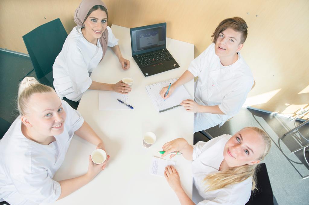 Fire unge mennesker med hvit uniform sitter sammen rundt et bord hvor de har gruppearbeid med skrivesaker og laptop. De ser smilende opp mot kamera. Foto.
