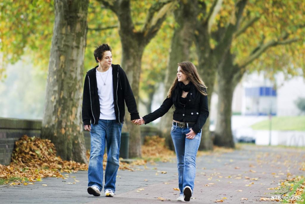 En tenåringsgutt og tenåringsjente på vei gjennom en park. De ser på hverandre mens de holder hverandre i hånden. Foto. 
