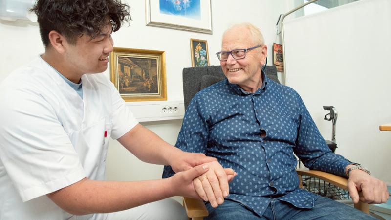 Ung mannlig helsefagarbeider sammen med en eldre mannlig pasient i blå skjorte. De smiler til hverandre. Foto.