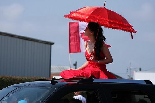 Lady dressed in red riding on the top of a car with an umbrella. Photo.