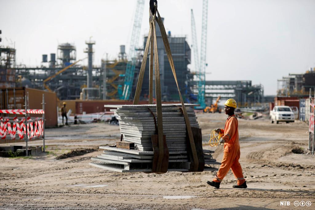Photo: In the foreground is a man in orange work clothes and a yellow construction helmet, carrying yellow wires. Behind him we see a pile of building materials and the outlines of an oil refinery. 