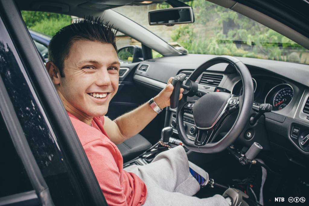 En mann med amputerte bein og arm kjører bil. Han ser mot kamera og smiler. Foto.
