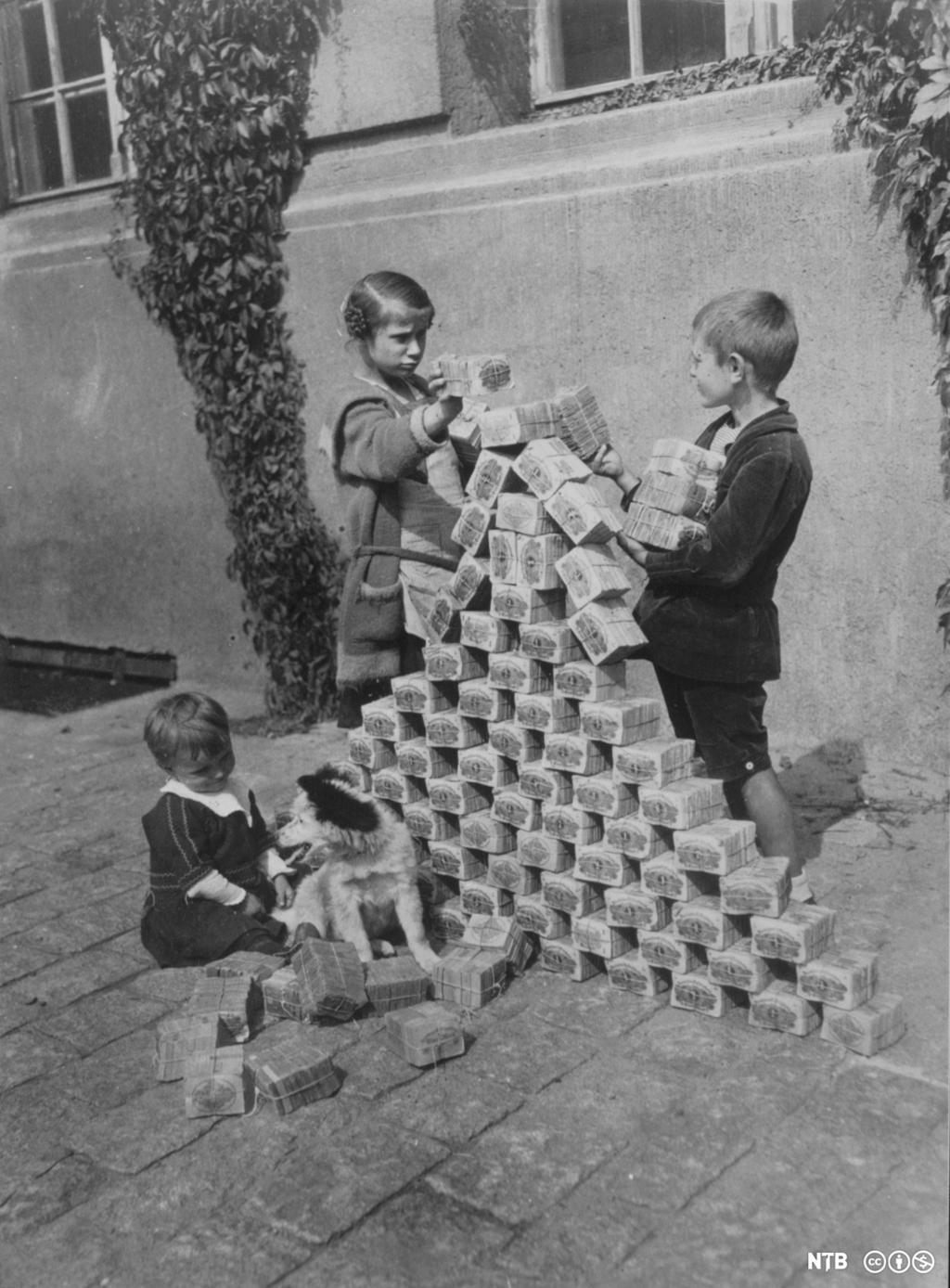 Tre barn leker med å stable seddelbunker i et tårn. Svart-hvitt-foto. 