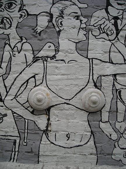 Veggmaleri av ei kvinne i bikini som blåser i ei fløyte. To store knotter på veggen utgjør puppene hennes. Foto. 