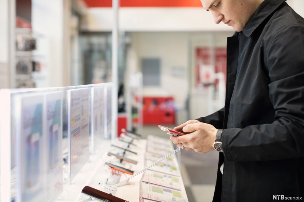 En ung mann står ved en rekke tyverisikrede mobiltelefoner i en butikk og ser nærmere på en av dem. Foto.