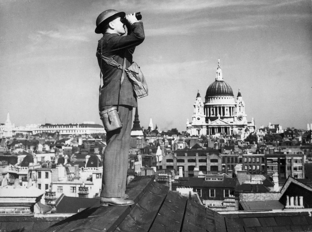We see a man with binoculars standing on a roof. In the background we see St. Paul's Cathedral and other buildings. Photo. 