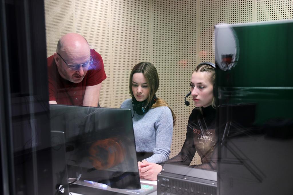 En lærer og to elever sitter foran en skjerm i et TV-kontrollrom. Det er gjenskinn med noe lys siden bildet er tatt gjennom glass. Foto.