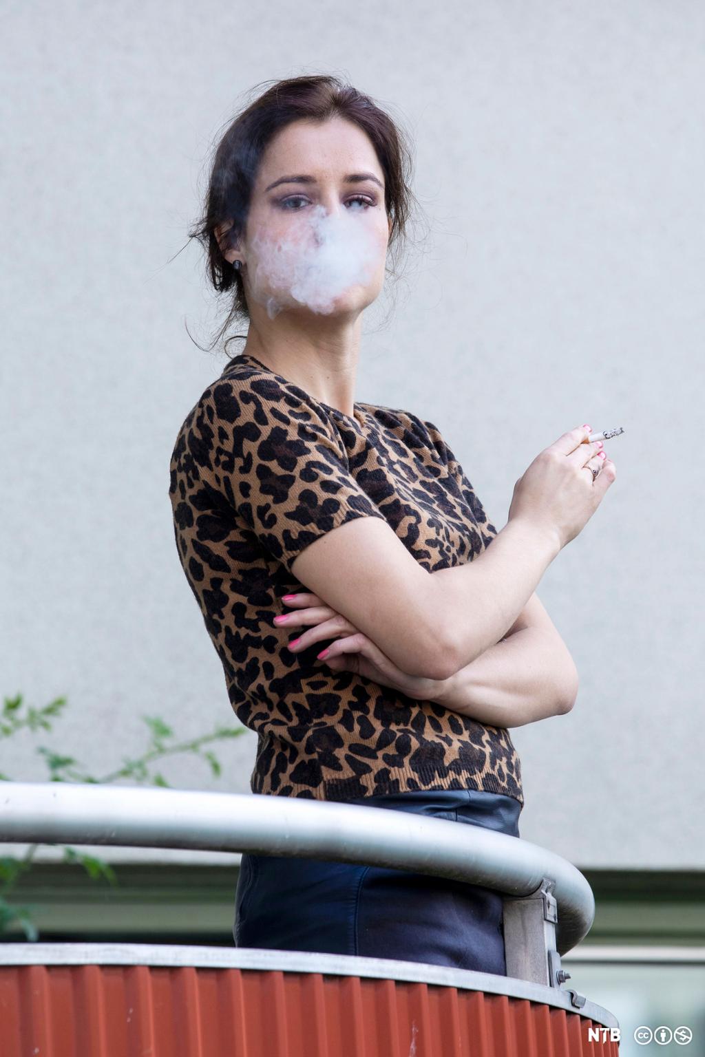 En kvinne står på balkongen og røyker. Hun blåser røyk mot fotografen. Foto.