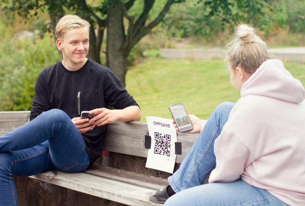 To unge mennesker med en smarttelefon på en parkbenk. Mellom dem er det et oppgaveark med en QR-kode. Foto.