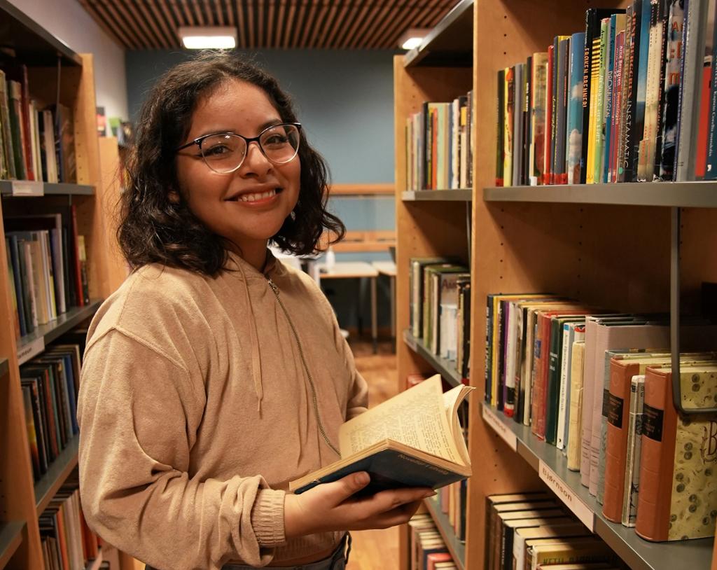 Ei jente står mellom hyller på eit bibliotek og smiler til kamera med ei open bok i hendene. Foto.