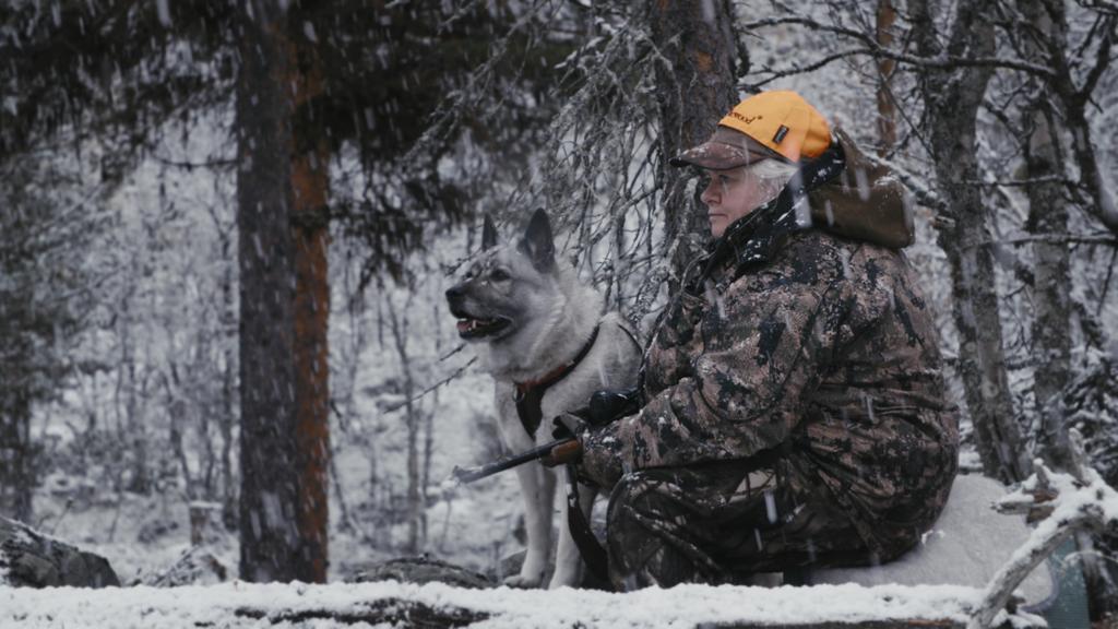 Foto av en eldre dame som holder ei rifle og sitter i en skog i snøvær. Ved siden av henne sitter en grå elghund. De ser i samme retning.