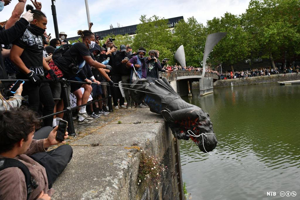 Mange bidrar og kaster den svære statuen ned i vannet. En stor menneskemengde står tett i tett på kaikanten, på en bro og langs kaikanten på den andre sida . Foto.