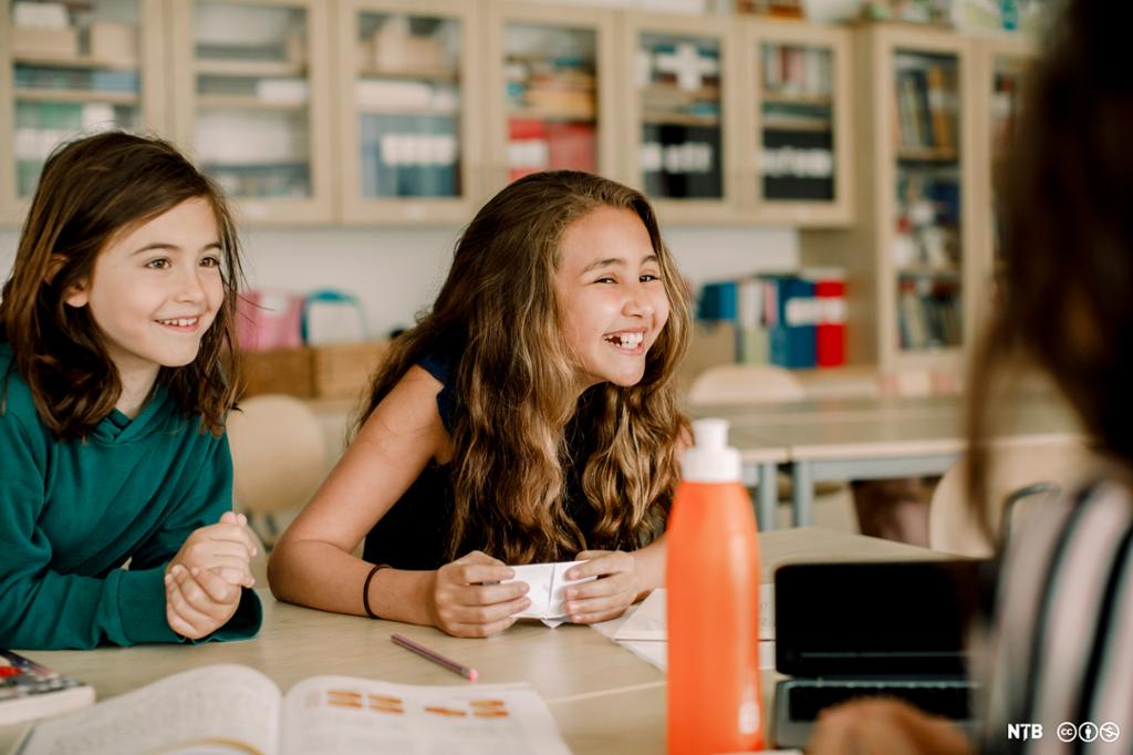 To smilende jenter sitter sammen i klasserommet. Foto.