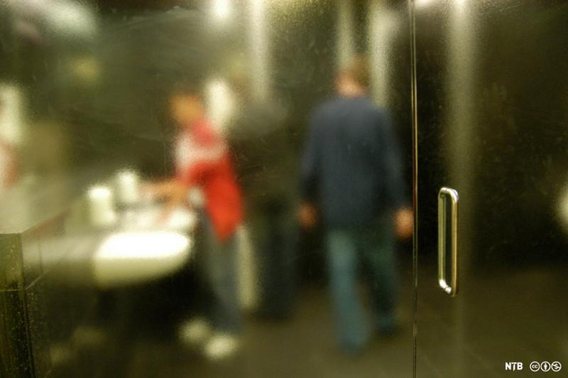 Uklart bilde tatt gjennom ei glassdør. To gutter er inne på et toalett. Den ene står og vasker hendene, og den andre går bak ham. Foto.