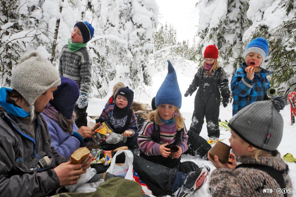 Barnehage har spisepause på skitur. Voksne og mange barn sitter i snøen og spiser, noen av barna står. Ei jente i bakgrunnen har på ski. Foto.