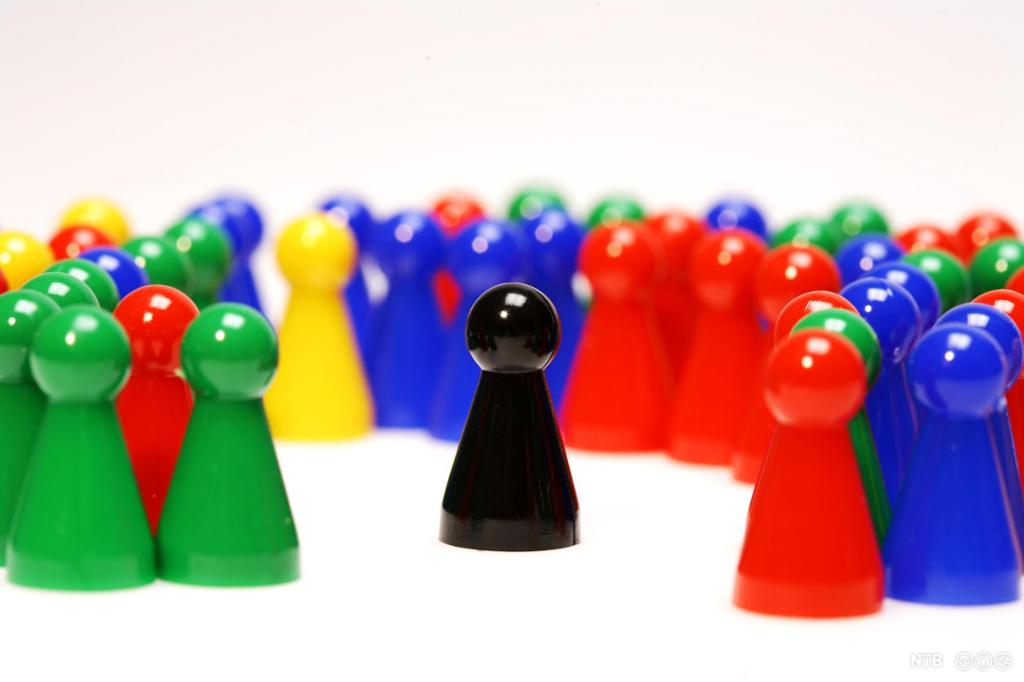 Game pieces in different colours. One black game piece is standing by itself. It is surrounded by the others, but there is a gap between it and them. Photo.