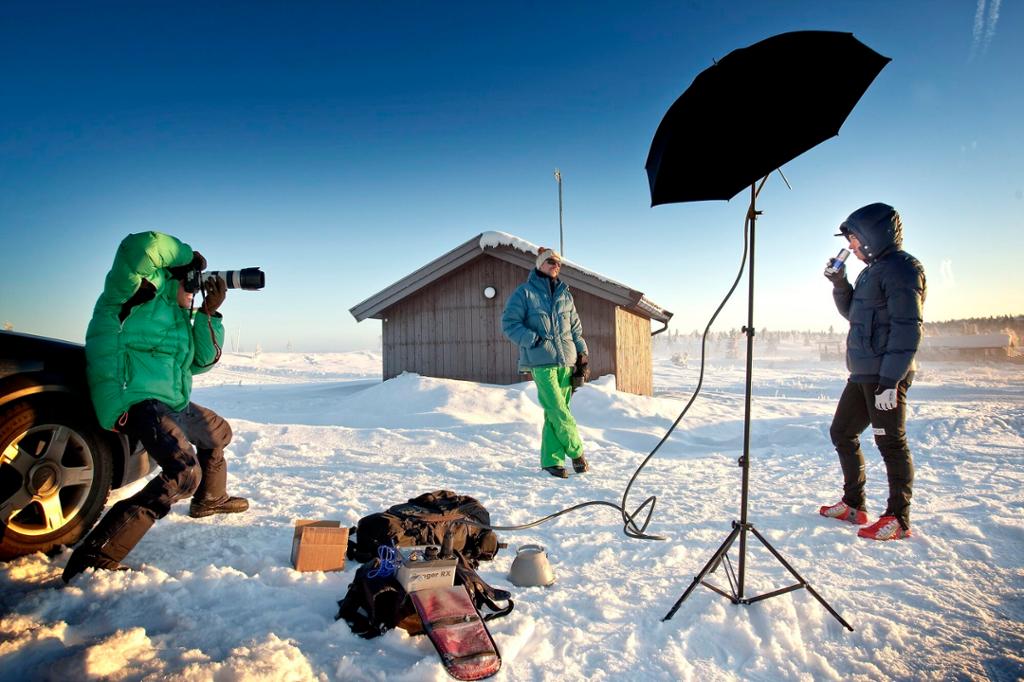 Mann i boblejakke står ute i snøen og drikker fra en drikkeboks mens han blir fotografert. Fotografen bruker en blitslampe med sølvparaply. Foto.