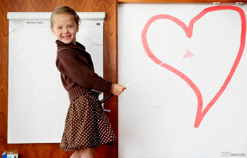 Smilende jente peker med pekestokk på et hjerte på ei tavle. Foto.