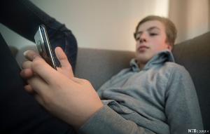 Gutt sitter med beina høyt i en sofa og sjekker mobilen sin. Foto.