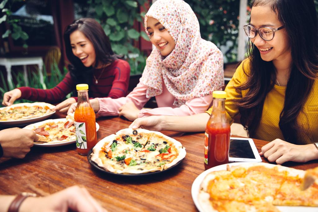Tre unge kvinner spiser pizza på en restaurant. En av dem bærer hijab. Foto.