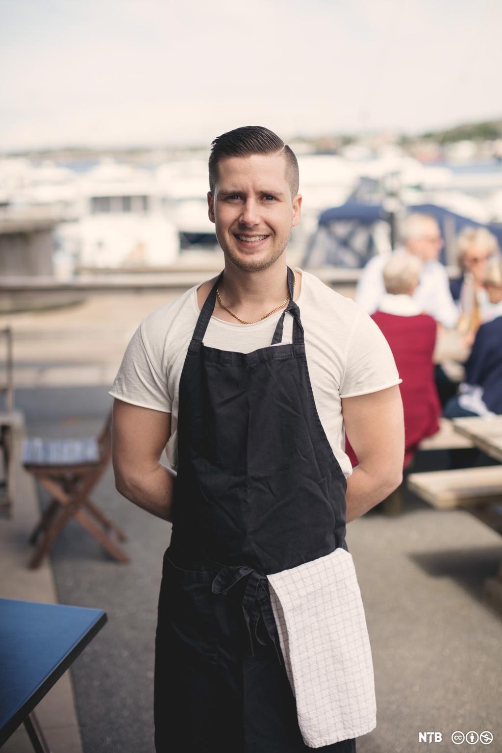Smilande, ung mannleg servitør med svart forkle på ein uterestaurant. Foto.
