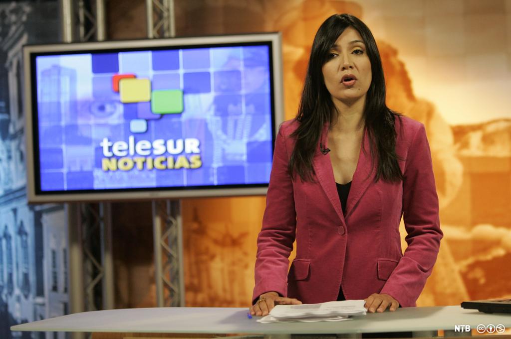 Et nyhetsanker, en ung kvinne med mørkt hår og rød jakke, snakker inn i et TV-kamera. I bakgrunnen ser vi en TV-skjerm med Telesurs logo. Foto.