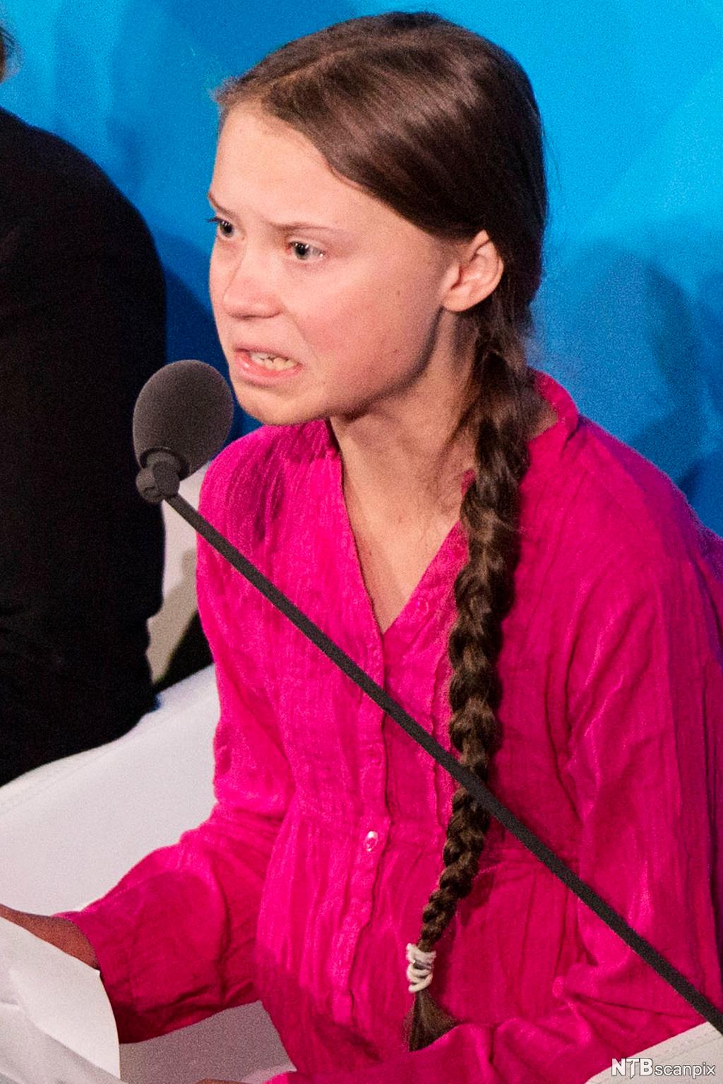 Greta Thunberg holder tale i FN. Ansiktsuttrykket hennes viser vrede og fortvilese over situasjonen hun taler om. Hun er kledd i rosa, og håret er fletta i ei lang flette.