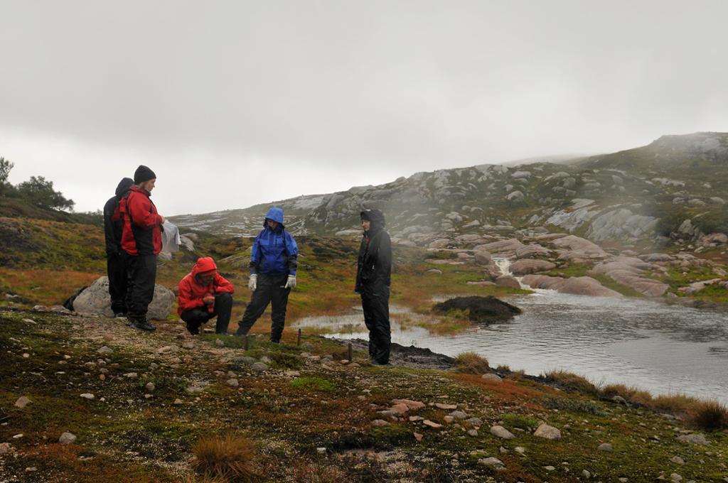 Personar i regntøy står i ring rundt person som sit på huk i fjellandskap. Foto.