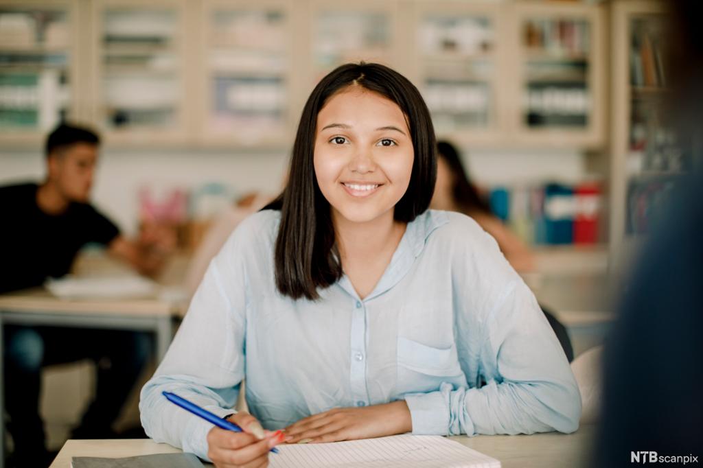 Ung, voksen elev sitter ved en pult og smiler. Hun har ei skriveblokk foran seg og en penn i hånda. Foto.