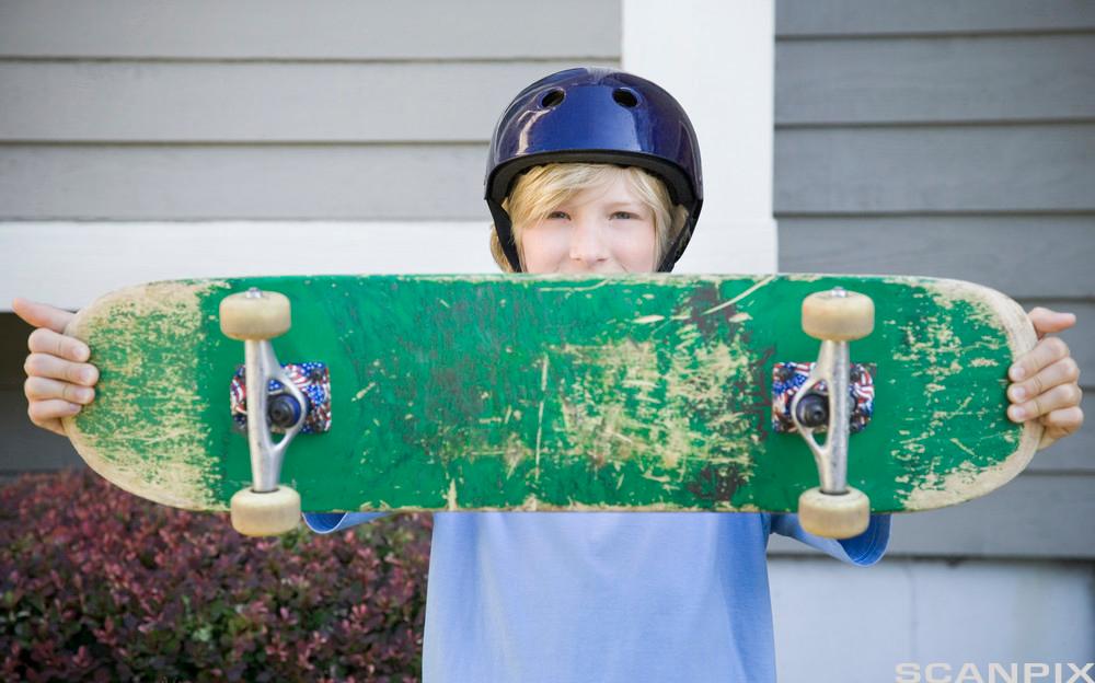 Gutt med hjelm på hodet holder opp et skateboard foran seg. Foto.