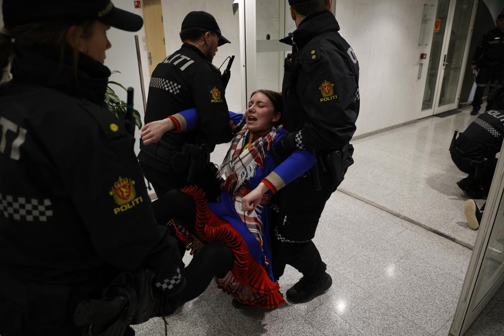 Politifolk bærer ei ung kvinne iført samekofte gjennom en korridor. Lenger nede i korridoren er det flere politifolk. Foto.