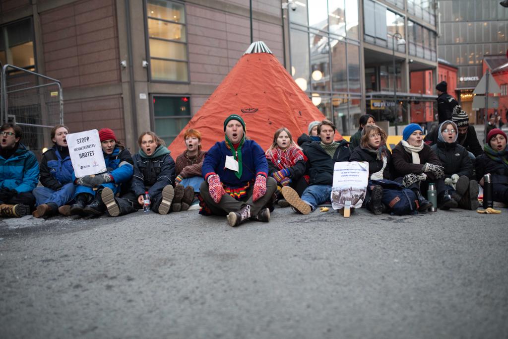 Ungdommer med plakater sitter i ei bygate foran en lavvo. Noen av ungdommene er iført samedrakt. Foto.