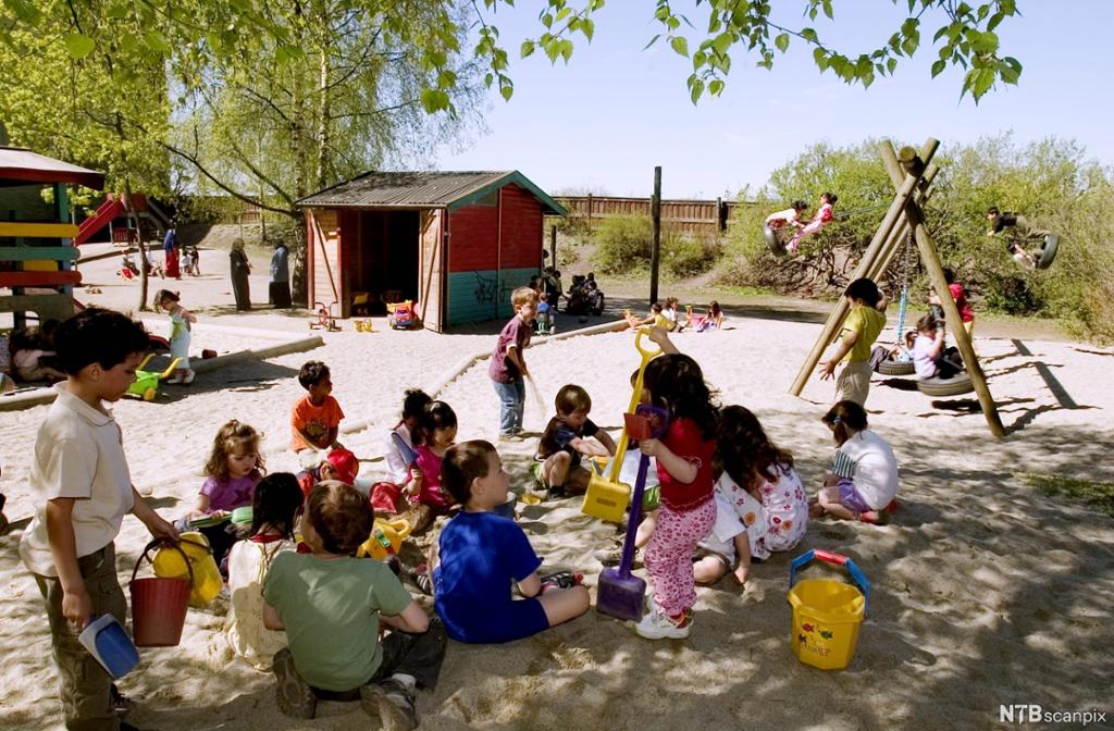 Mange barn leker ute i en barnehage. Noen leker i sandkassa, noen andre husker, og noen gjør andre ting. Det er sol, og mange av barna er i skyggen. Foto.