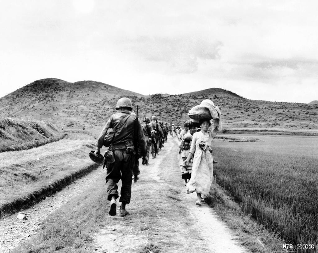 En rekke med soldater passerer en rekke med kvinner og barn som bærer eiendeler på hodet. Svart-hvitt foto. 